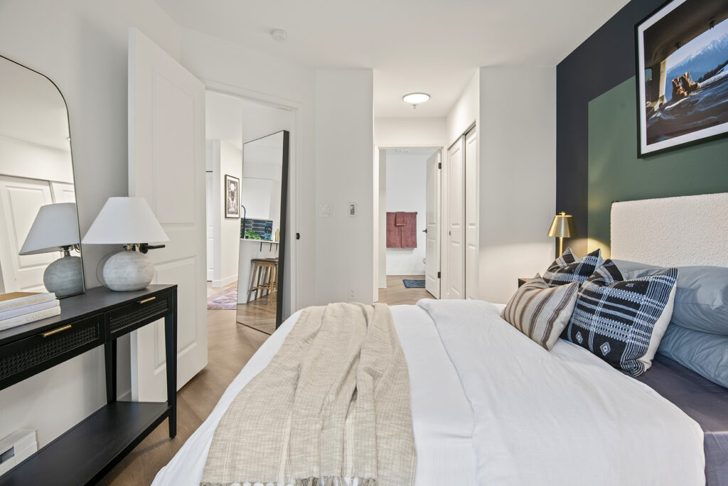 A bedroom with a bed and a mirror. The bed has a comforter and pillows. The mirror is on the wall behind the bed.