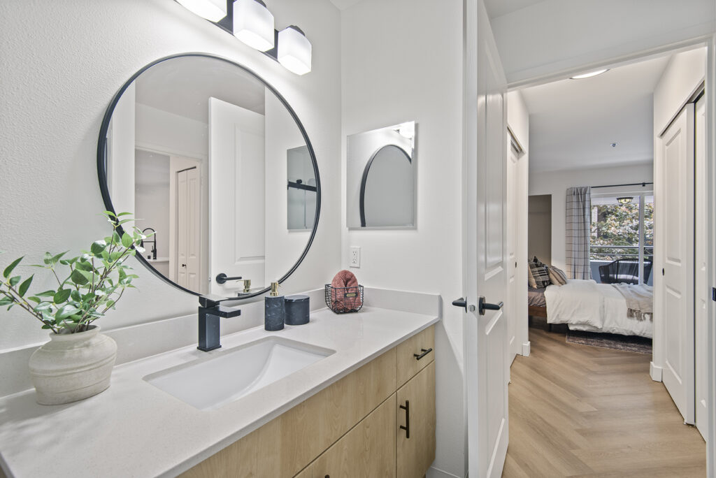 A bathroom with a round mirror and a pedestal sink. The sink has a faucet and a drain. The bathroom is next to a bedroom. There is a doorway to the bedroom in the image.