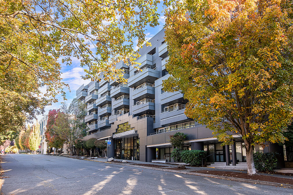 A modern mid-rise apartment building with balconies, surrounded by trees with green and autumn-colored leaves, lines a quiet, empty street under a partly cloudy sky.
