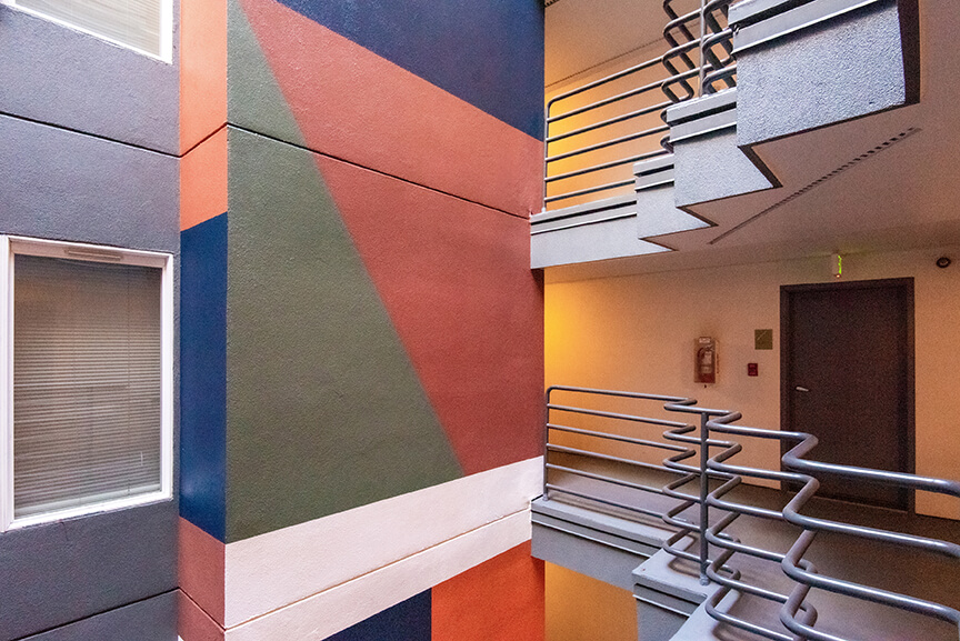 A modern indoor staircase with metal railings and colorful geometric patterns painted on the adjacent wall, featuring shades of blue, green, orange, and white. There are doors and windows on each floor.