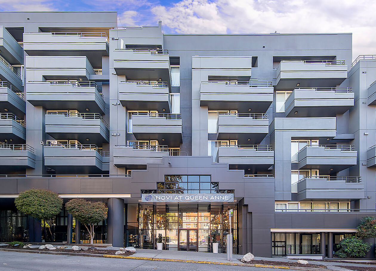 Modern multi-story apartment building with numerous balconies and large windows, displaying a sleek gray exterior. The entrance is glass-fronted with the sign NOVI AT QUEEN ANNE above the doors. Trees flank the building.