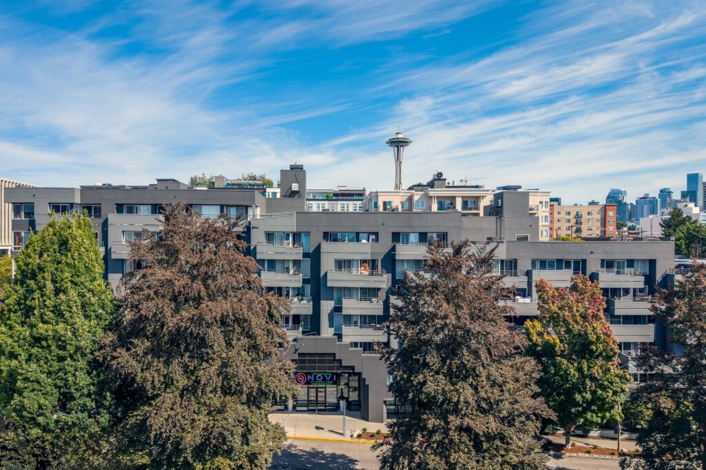 A modern gray apartment building is surrounded by tall trees, with the Space Needle and part of the Seattle skyline visible in the background under a blue sky with wispy clouds.