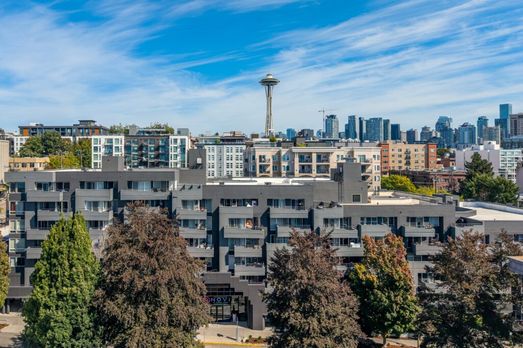 Apartment buildings and trees with the Seattle skyline and Space Needle in the background under a blue sky with wispy clouds.