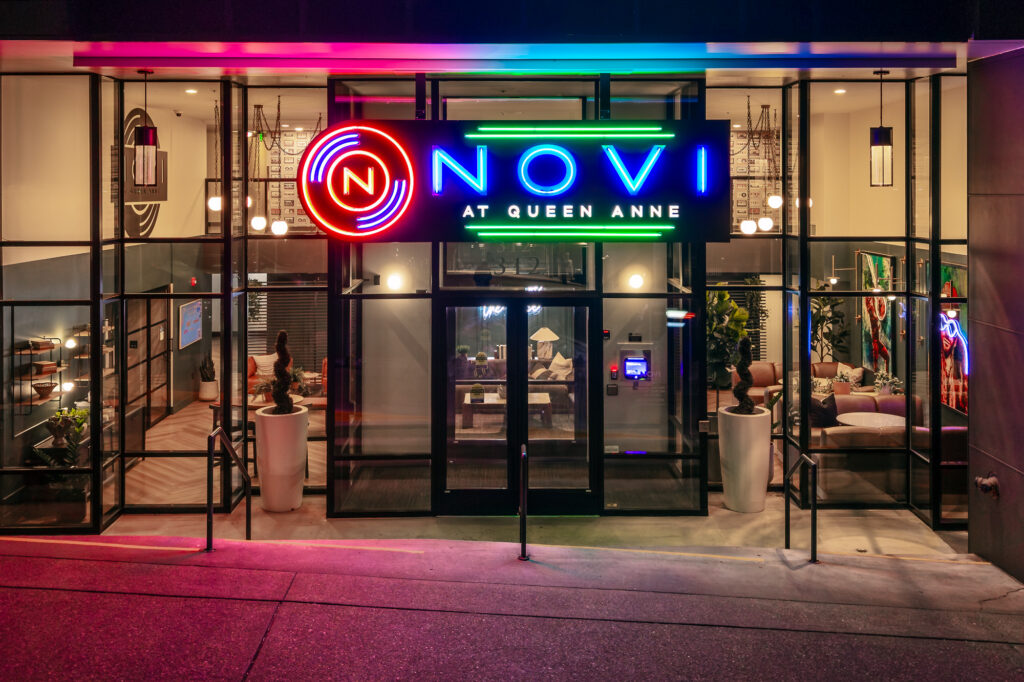 Neon sign reading NOVI AT QUEEN ANNE above glass doors of a modern, well-lit building lobby with decor, couches, and plants visible inside.