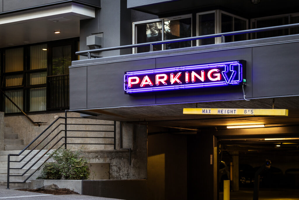A brightly lit neon PARKING sign above the entrance to an underground garage. A yellow bar marked MAX HEIGHT 65 hangs from the ceiling. Stairs and railings are visible to the left.