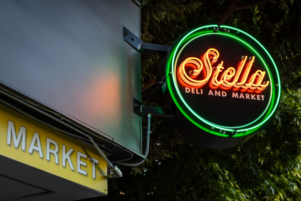 A round neon sign reads Stella Deli and Market in yellow and red, bordered by green, above a storefront with a yellow MARKET sign. Trees are in the background.