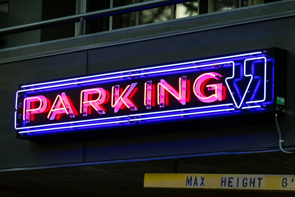 A neon sign with the word PARKING in red and a blue outline arrow pointing right. The sign is mounted above a parking garage entrance. A yellow bar below reads MAX HEIGHT 67..