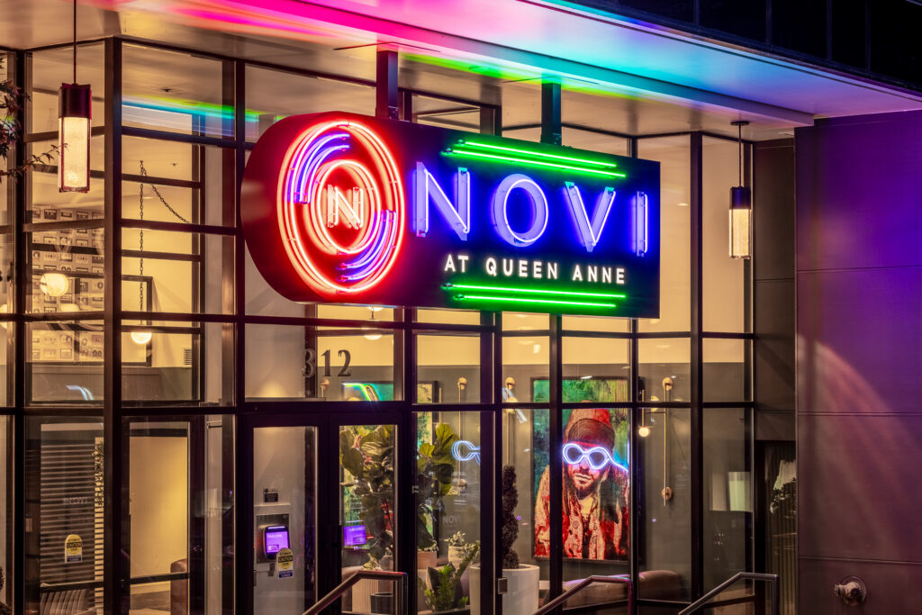 Colorful neon sign reads Novi at Queen Anne above glass doors. The interior has visible plants and a vibrant portrait of a person with bright lights and glasses. Address number 312 is on the entrance.